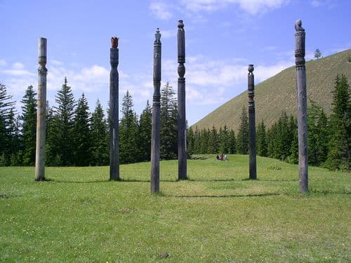 Tuymaada Valley (Yakutsk) Location: Russia, Saha, Markha Coordinates: 62.0812219, 129.763012 Caption: Seven tall, weathered wooden poles stand in a grassy field under a blue sky, arranged in a loose circle. Each pole features carved tops, with one displaying a reddish-brown cap. A dense evergreen forest borders the left side, while a rolling green hill rises on the right. In the distance, two small figures sit near the center, dwarfed by the monument-like structure. The scene evokes an ancient c